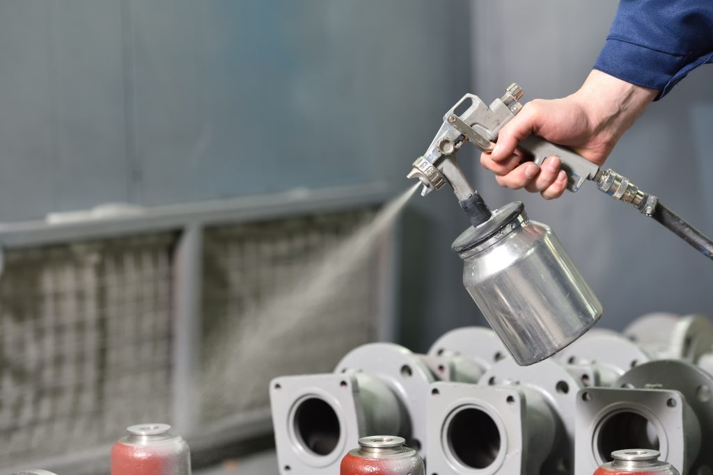 A worker at a factory in a special room paints parts of the valves from the spray gun. Paint valves in gray color, hand close up
